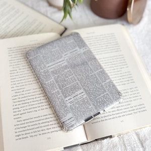 May include: A rectangular e-reader case featuring a vintage newspaper print with black text on a light gray background. The case has a slim profile with a clear plastic edge and a small metal ring attachment at the bottom center for accessories.