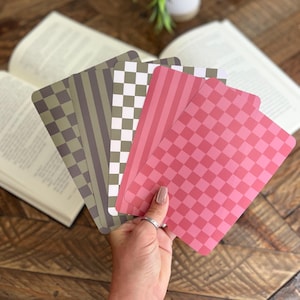 May include: A hand holds a fan of patterned cards in shades of pink, green, and gray. The patterns include stripes, checks, and a grid design. Open books and a small potted plant are in the background on a wooden surface.