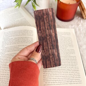 Op de afbeelding: Een bruine boekenlegger met een houtnerfpatroon wordt boven een open boek gehouden. De boekenlegger heeft een rechthoekige vorm met verticale lijnen, die houten planken nabootsen. Een kaars en een plant zijn zichtbaar op de achtergrond.