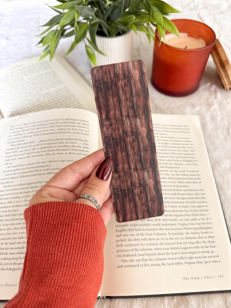 Op de afbeelding: Een bruine boekenlegger met een houtnerfpatroon wordt boven een open boek gehouden. De boekenlegger heeft een rechthoekige vorm met verticale lijnen, die houten planken nabootsen. Een kaars en een plant zijn zichtbaar op de achtergrond.