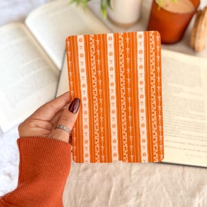 Pu&ograve; includere: Una cover rettangolare per e-reader con un motivo a righe verticali nei toni dell'arancione e del bianco. Il design presenta motivi ripetuti di piccole zucche, fiocchi delicati e minuscole foglie disposte in colonne alternate.