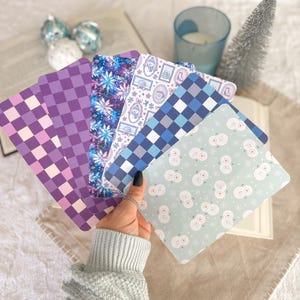 May include: A set of five rectangular bookmarks featuring diverse patterns including purple and light pink checkers, a dark blue floral design, a white background with small winter icons, a blue and grey plaid, and a light green snowman print.