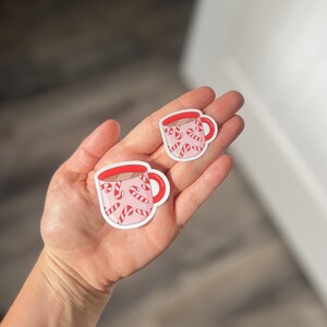 May include: Two pink and red mugs with candy cane designs. The mugs are on a white background.