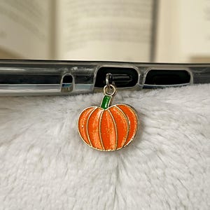 May include: A small, orange pumpkin charm with a gold outline and green stem. The charm is attached to a phone case. The pumpkin is a fall-themed accessory.