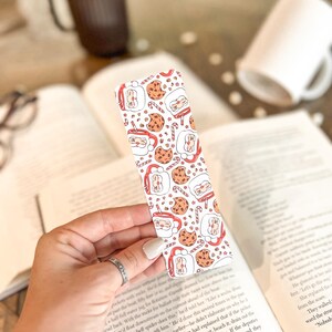 May include: White bookmark with a repeating pattern of Santa Claus, cookies, and candy canes. The bookmark is being held by a hand.