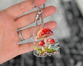 May include: A silver key chain with a charm featuring a cluster of red and white mushrooms with white spots, surrounded by green foliage.