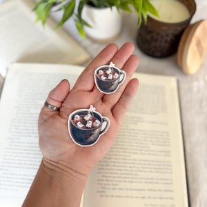 May include: Two stickers featuring a black mug filled with white ghost marshmallows. The stickers are held in a hand, with a book and a candle in the background. The stickers are perfect for Halloween or spooky season.
