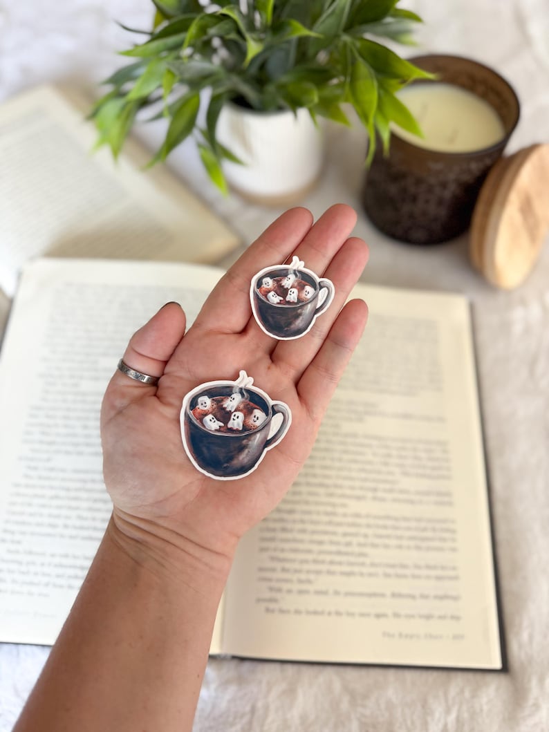 May include: Two stickers featuring a black mug filled with white ghost marshmallows. The stickers are held in a hand, with a book and a candle in the background. The stickers are perfect for Halloween or spooky season.