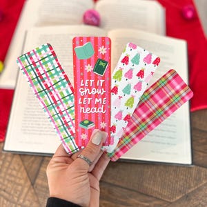 May include: Four Christmas-themed bookmarks. One bookmark features a plaid pattern in pink and green. Another bookmark has a white background with pink and green Christmas trees. A third bookmark has a pink and white striped background with a green book and the text "Let it snow let me read". The fourth bookmark has a pink and green plaid pattern.