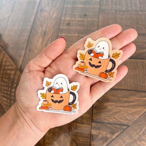 May include: Two stickers featuring a cartoon ghost reading a book inside a jack-o'-lantern mug. The mug is orange with a black handle and a carved face. There are small pumpkins, books, and fall leaves around the mug.