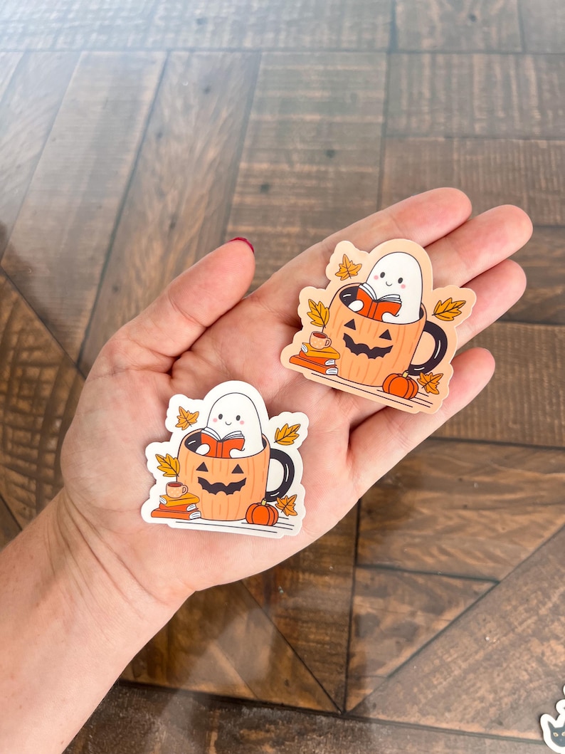 May include: Two stickers featuring a cartoon ghost reading a book inside a jack-o'-lantern mug. The mug is orange with a black handle and a carved face. There are small pumpkins, books, and fall leaves around the mug.