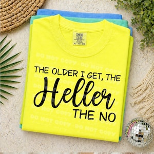 Puede incluir: Una pila de tres camisetas en amarillo, verde y azul. La camiseta amarilla superior tiene el texto negro "THE OLDER I GET, THE HELLER THE NO". La camiseta tiene una etiqueta que dice "COMFORT COLORS XL-TG-EG".