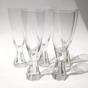 Könnte beinhalten: Ein Set aus fünf klaren Champagnergläsern mit hohen, schlanken Kelchen, die vertikale Ätzlinien aufweisen, sowie dicken, facettierten sechseckigen Füßen. Die Gläser sind in einer Gruppe angeordnet und zeigen ihre transparente, geometrische Form.