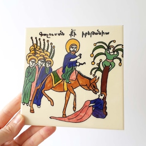 Könnte beinhalten: Eine quadratische Keramikfliese mit einer handgemalten Illustration einer Figur auf einem braunen Esel, begleitet von Anhängern und einem Baum. Die Szene zeigt eine Person, die ein rosa Tuch auf den Boden legt, und armenischen Text oben.