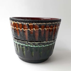 Puede incluir: Un cuenco de cerámica con exterior texturizado. El cuenco presenta un borde marrón oscuro, una sección media con un patrón marrón y naranja y una sección inferior verde oscuro. El interior es de color rojo intenso.