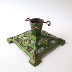 May include: A green metal Christmas tree stand with a square base and decorative leaf and dot patterns. The stand has a central cup with handles for holding the tree trunk. The stand is painted green with gold accents.