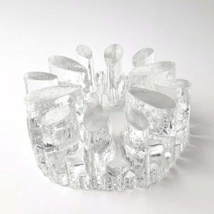 Op de afbeelding: Decoratieve kom van helder glas met een textuurrijk, organisch ontwerp. De kom heeft een cirkelvormige basis met talrijke rechtopstaande, cilindrische elementen, wat een unieke, artistieke uitstraling creëert. Het glas heeft een matte, ijzige uitstraling.