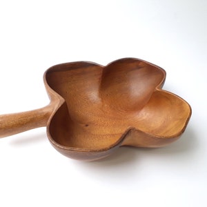 Pode incluir: Uma tigela de servir de madeira em forma de trevo de quatro folhas com uma alça. A tigela é feita de madeira marrom claro e tem um acabamento natural.