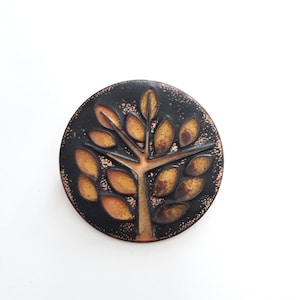 Könnte beinhalten: Eine runde Brosche in Schwarz und Kupfer mit einem stilisierten Baumdesign. Der Baum hat einen braunen Stamm und gelb-orange Blätter.