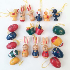 Puede incluir: Una colección de adornos de Pascua, que incluye conejitos de madera, pájaros y huevos pintados. Los adornos son de varios colores, como rojo, azul, amarillo y verde, y están ensartados con cordones amarillos. Los adornos están dispuestos sobre una superficie blanca.