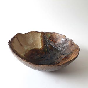 Può includere: Una ciotola in ceramica fatta a mano con una forma organica unica. La ciotola presenta una miscela di toni terrosi, tra cui marrone, beige e tocchi di verde. La superficie strutturata aggiunge interesse visivo, rendendola un pezzo decorativo.