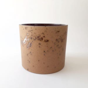 Puede incluir: Una maceta cilíndrica de cerámica con un exterior de color tostado mate que presenta un patrón de esmalte marrón moteado. El interior del recipiente tiene un acabado de esmalte marrón oscuro y liso, contrastando con la superficie exterior texturizada.