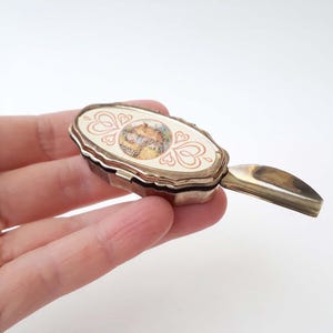 Könnte beinhalten: Ein ovales, antikes silberfarbenes Kompaktpuder mit einem Klappdeckel und einem kleinen, angebrachten Spiegel. Der Deckel hat ein cremefarbenes Emaille-Design mit einem zentralen Bild und herzförmigen Akzenten. Das Kompaktpuder ist geöffnet.