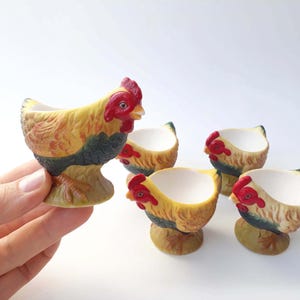 Può includere: Un set di cinque portauova in ceramica a forma di gallina. I portauova sono dipinti con colori giallo, rosso e marrone. Un portauovo è tenuto in mano. Gli altri quattro portauova sono su una superficie bianca.