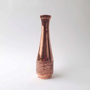 Pode incluir: Um vaso de cobre com um design texturizado. O vaso é alto e fino com uma abertura estreita.