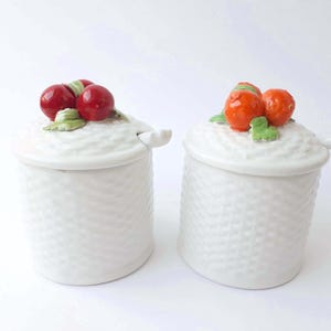 Può includere: Due zuccheriere in ceramica bianca con coperchi e cucchiaini. Una ciotola ha un design di ciliegie rosse e l'altra ha un design di agrumi arancioni.