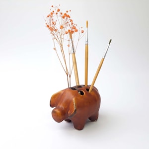 Könnte beinhalten: Ein brauner, keramik-geformter Stiftehalter in Form eines Schweins mit drei Pinseln und getrockneten Blumen, die aus den Löchern herausragen.