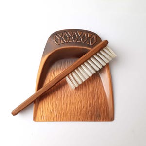 Könnte beinhalten: Ein Vintage-Set aus kupferfarbener Kehrschaufel und Bürste. Die Kehrschaufel hat eine strukturierte Oberfläche und ein dekoratives geometrisches Muster oben. Die Bürste hat einen Holzgriff und weiße Borsten. Ein klassisches Reinigungswerkzeug.