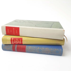 Könnte beinhalten: Ein Stapel von drei alten gebundenen Büchern mit verschiedenen farbigen Einbänden. Das oberste Buch hat einen weißen Einband mit dem Text "Liebe in unserer Zeit" in Rot. Das mittlere Buch hat einen gelben Einband mit dem Text "Zwei Töchter auf Pump" in Schwarz. Das unterste Buch hat einen blauen Einband mit goldenen Lettern.