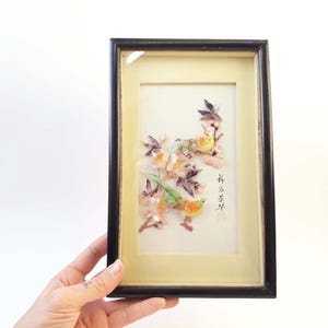 May include: Framed artwork featuring two birds perched on flowering branches. The birds have yellow, green, and orange plumage. The artwork is set against a cream-colored background within a black frame. The image includes Asian characters.