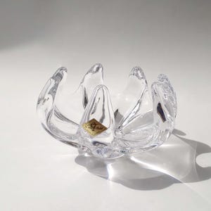 Könnte beinhalten: Klarer Glasbehälter mit einem einzigartigen, blütenblattartigen Design. Die Schale hat eine breite, offene Form mit mehreren gebogenen Kanten. Ein kleines, goldfarbenes Etikett ist vorne angebracht. Der Behälter steht auf einer weißen Oberfläche.