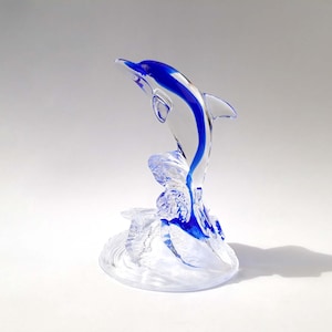 Puede incluir: Una escultura de delfín de cristal transparente con una vibrante franja azul en la espalda y la cola. El delfín salta desde una base en forma de ola, creando una pieza decorativa dinámica y elegante. Una hermosa obra de arte en vidrio.