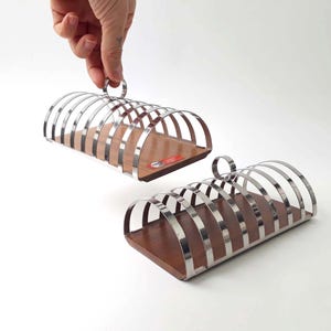 Può includere: Due portatovaglioli vintage in cromo e legno. Ogni portatovagliolo presenta barre cromate curve e una base in legno. Uno dei portatovaglioli è tenuto in mano. Progettati per contenere fette di pane tostato in posizione verticale.