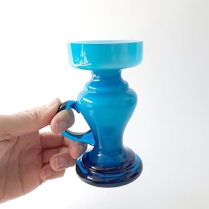 Könnte beinhalten: Eine blaue Glasvase mit Henkel. Die Vase hat eine breite Öffnung und einen schmalen Boden. Die Vase ist aus dickem, undurchsichtigem Glas.