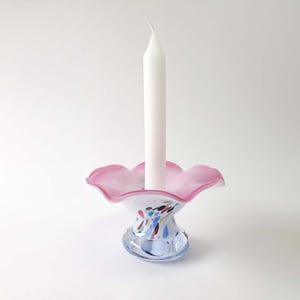 Può includere: Una candela bianca è inserita in un portacandele decorativo in vetro. Il portacandele ha un bordo rosa e increspato e una base blu con macchie colorate. Un elemento decorativo per la casa.