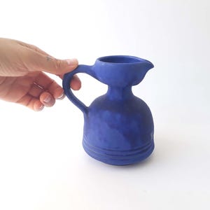 Peut inclure: Une cruche en céramique bleu cobalt avec une anse et un bec verseur. La cruche a une base bulbeuse et un col cylindrique. La poignée est arquée et le bec verseur est petit et incliné. La cruche est tenue par une main.