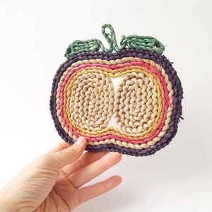 Può includere: Un sottopentola intrecciato a forma di mela con un arcobaleno di colori, tra cui rosa, arancio, giallo e verde. Il sottopentola è realizzato in fibre naturali e ha un bordo esterno viola.