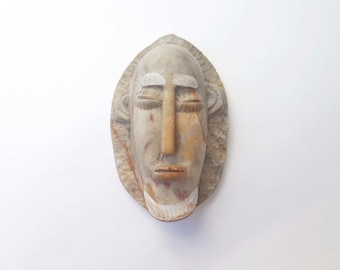 Pode incluir: Uma máscara de madeira esculpida com um rosto humano estilizado. A máscara é principalmente bege com detalhes em castanho e vermelho. O rosto tem olhos fechados, um nariz longo e uma boca pequena. A máscara é oval e parece ser uma obra de arte tribal africana.