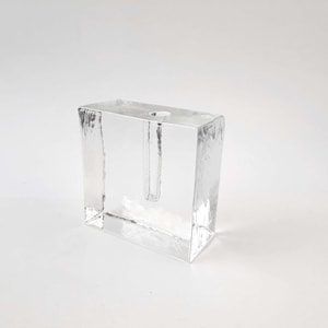 Könnte beinhalten: Eine klare, rechteckige Glasvase mit strukturierter Oberfläche. Die Vase hat eine einzelne, schmale Öffnung für einen Stiel. Das Design ist minimalistisch und modern, geeignet für eine einzelne Blume oder ein kleines Arrangement.