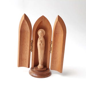 Pode incluir: Uma figura religiosa de madeira de uma mulher dentro de um tríptico de madeira. A figura é castanho claro, e o tríptico tem três painéis arqueados que se abrem sobre dobradiças de latão. A base é castanho mais escuro e o fundo é branco.