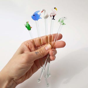 Peut inclure: Ensemble de bâtonnets à cocktail avec des figurines d'animaux. Les figurines comprennent un poisson bleu, un cygne blanc, un flamant rose, une grenouille verte et un dauphin vert. Les bâtonnets sont transparents et tenus dans une main.