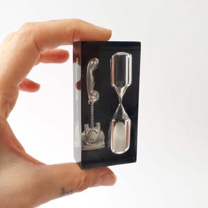 Może przedstawiać: Czarny, prostokątny element dekoracyjny z srebrnym telefonem i przezroczystą klepsydrą. Klepsydra ma w środku biały piasek. Telefon ma styl vintage. Element jest trzymany przez dłoń.