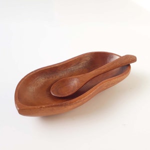 Puede incluir: Un plato de servir de madera con forma de cacahuete con una cuchara de madera a juego. El plato es de color marrón oscuro y la cuchara es ligeramente más clara.