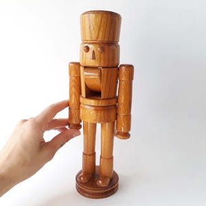 Könnte beinhalten: Holz-Nussknacker-Figur mit hellbraunem Finish. Die Figur hat ein traditionelles Design mit hohem Hut, geschnitztem Gesicht und beweglichen Armen und Beinen. Der Nussknacker steht auf einem runden Sockel.