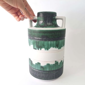 May include: A ceramic vase with a cylindrical shape, featuring a dark green top and base, with a white band in the middle. The vase has two small handles and is decorated with a dripping green paint effect. The vase is approximately 30 cm tall.