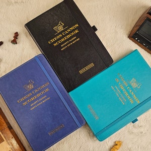 May include: Three blue, black, and turquoise hardcover notebooks with gold lettering. The notebooks are titled "Chess Cannon Scorebook" and feature a chess piece graphic.